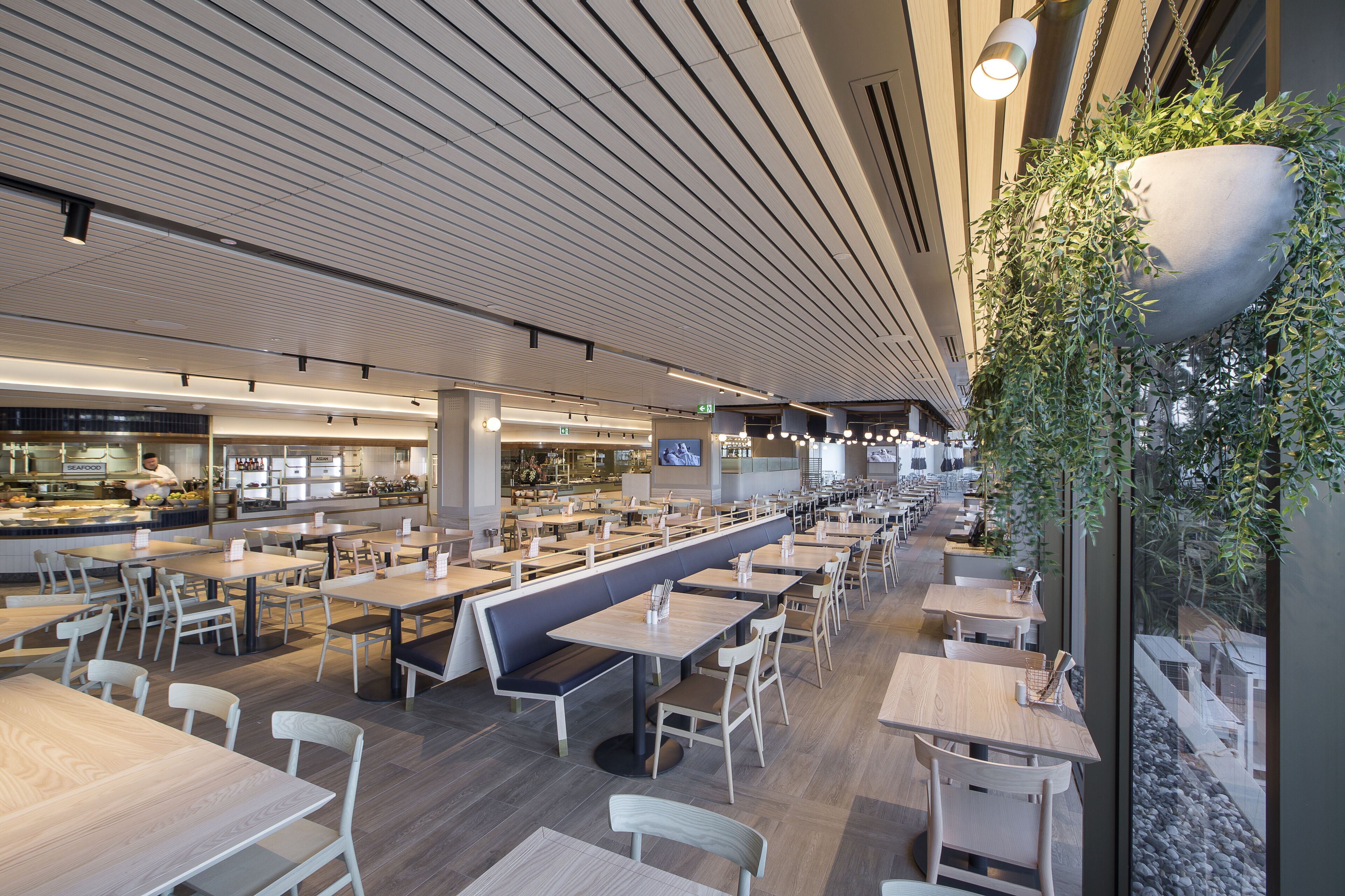 Une cafétéria spacieuse avec de longues tables communes et des places assises individuelles, un design de plafond moderne et des jardinières suspendues qui apportent une touche de verdure.