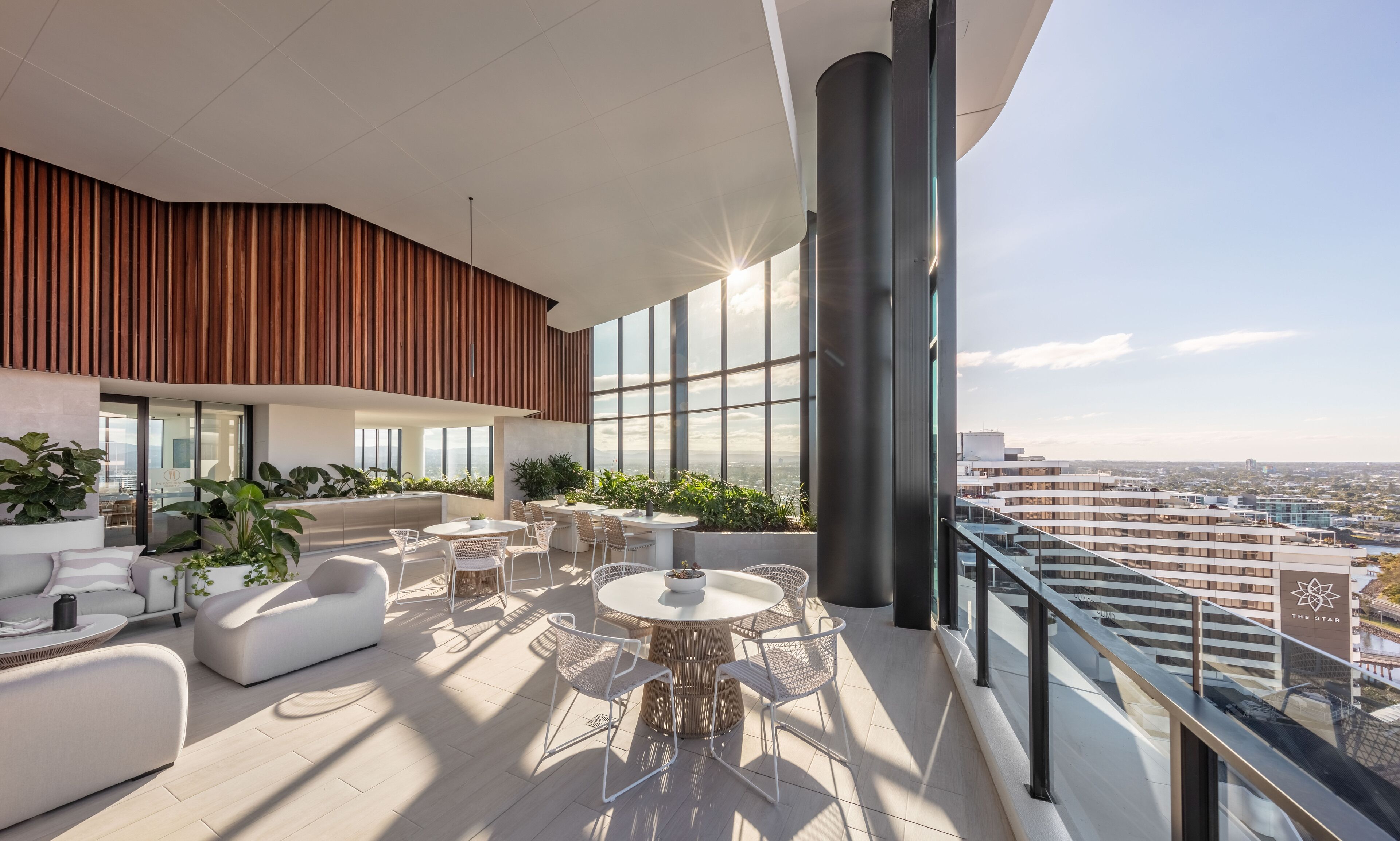 A modern rooftop lounge with a panoramic city view, featuring a round dining table, white sofas, and potted plants.