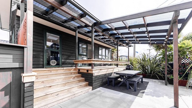 A covered patio with a wooden bench and table, surrounded by potted plants. - Manaia Magic - Tairua Holiday Home (Tairua)