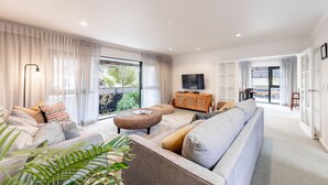 A modern living room with a sofa, ottoman, and a television on a wooden cabinet. - Manaia Magic - Tairua Holiday Home (Tairua)