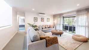 A modern living room with a grey sofa, a patterned ottoman, and a large window with sheer curtains. - Manaia Magic - Tairua Holiday Home (Tairua)