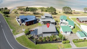 An aerial view of a residential area with houses, a road, and a beach in the background. - Manaia Magic - Tairua Holiday Home (Tairua)