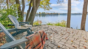 A wooden deck with a bench and a plaid blanket, overlooking a calm lake with trees and distant land.