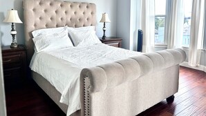 A bedroom with a tufted headboard, two bedside lamps, a large window with curtains, and a wooden floor.