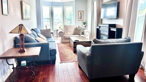 A modern living room with a sofa, armchairs, a coffee table, and a TV. There are large windows with curtains, a lamp, and framed pictures on the walls.