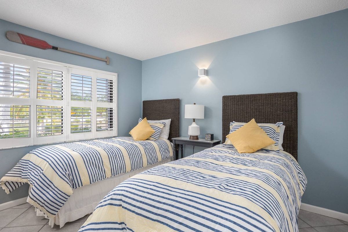 A room with two beds, a window with blinds, a wall-mounted lamp, and a paddle on the ceiling.