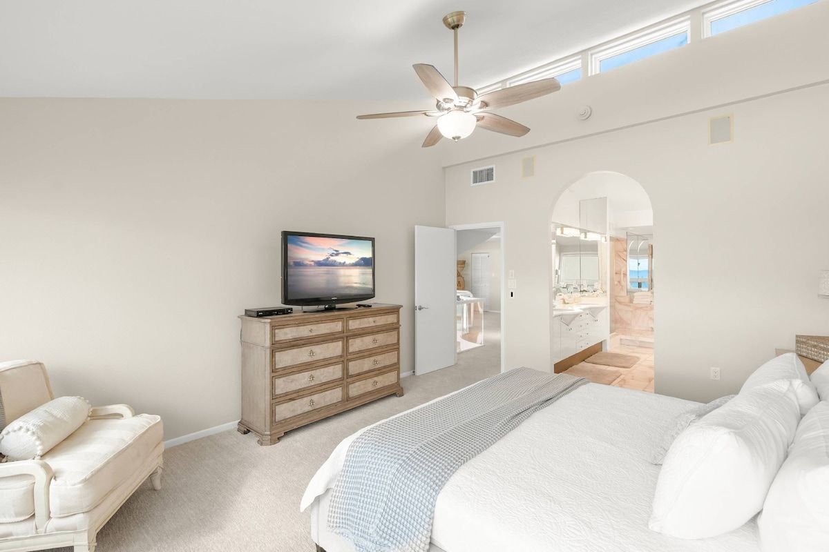 A bedroom with a bed, a television on a dresser, a ceiling fan, and an open door leading to a bathroom.