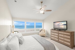 A bedroom with a bed, a television on a dresser, a chair, and a view of the ocean through a large window.
