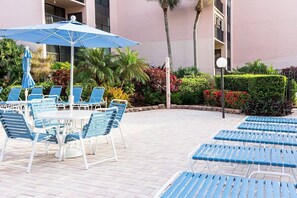 A poolside area with lounge chairs and a table under a blue umbrella, surrounded by greenery and a building facade. - Unbeatable Ocean Views | Private Beach | Penthouse (Sarasota)
