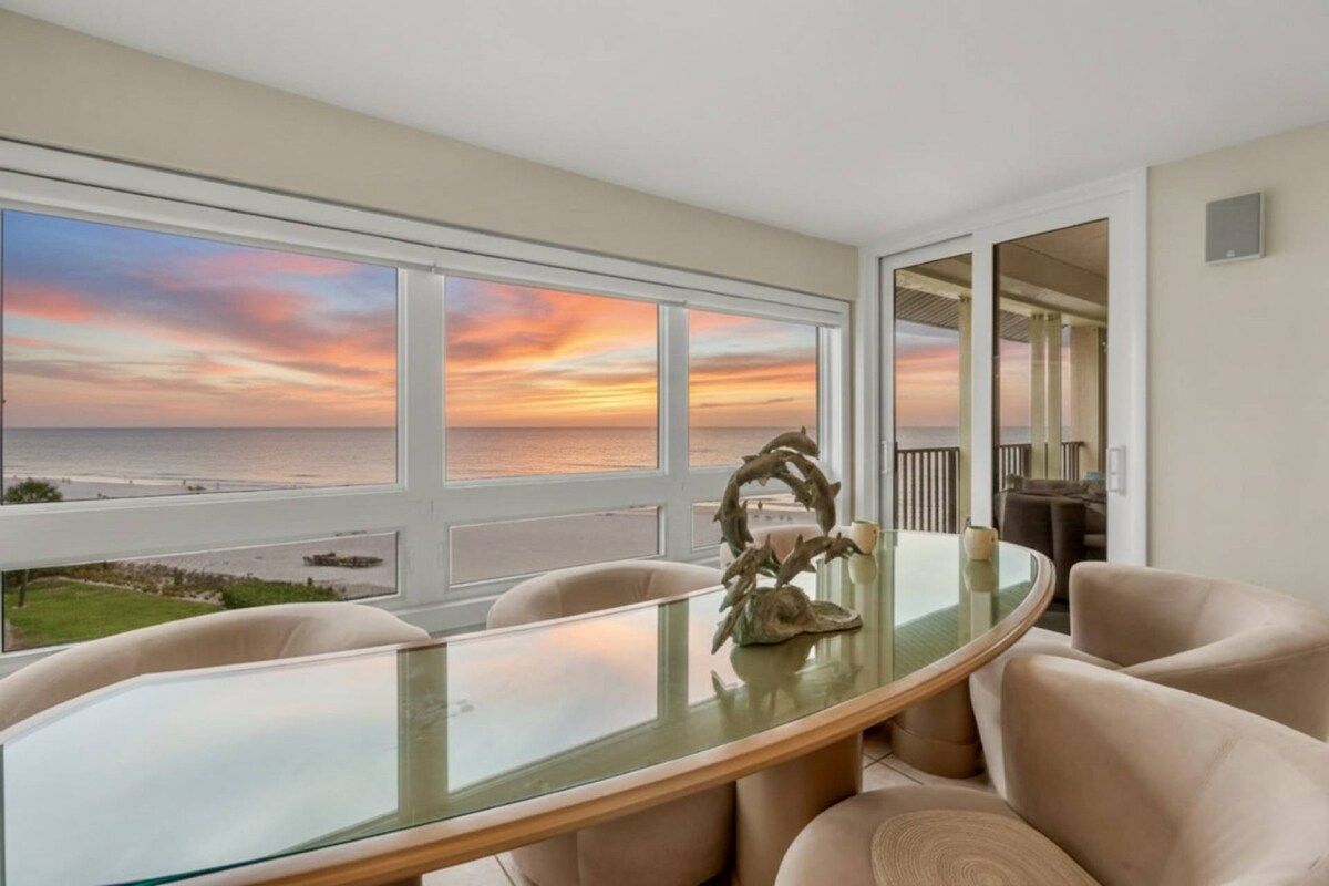 A dining area with a glass table, beige chairs, and a view of the ocean at sunset.