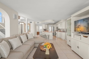 A modern living room with a sofa, coffee table, and a TV. There is a kitchen in the background with a bar and stools.