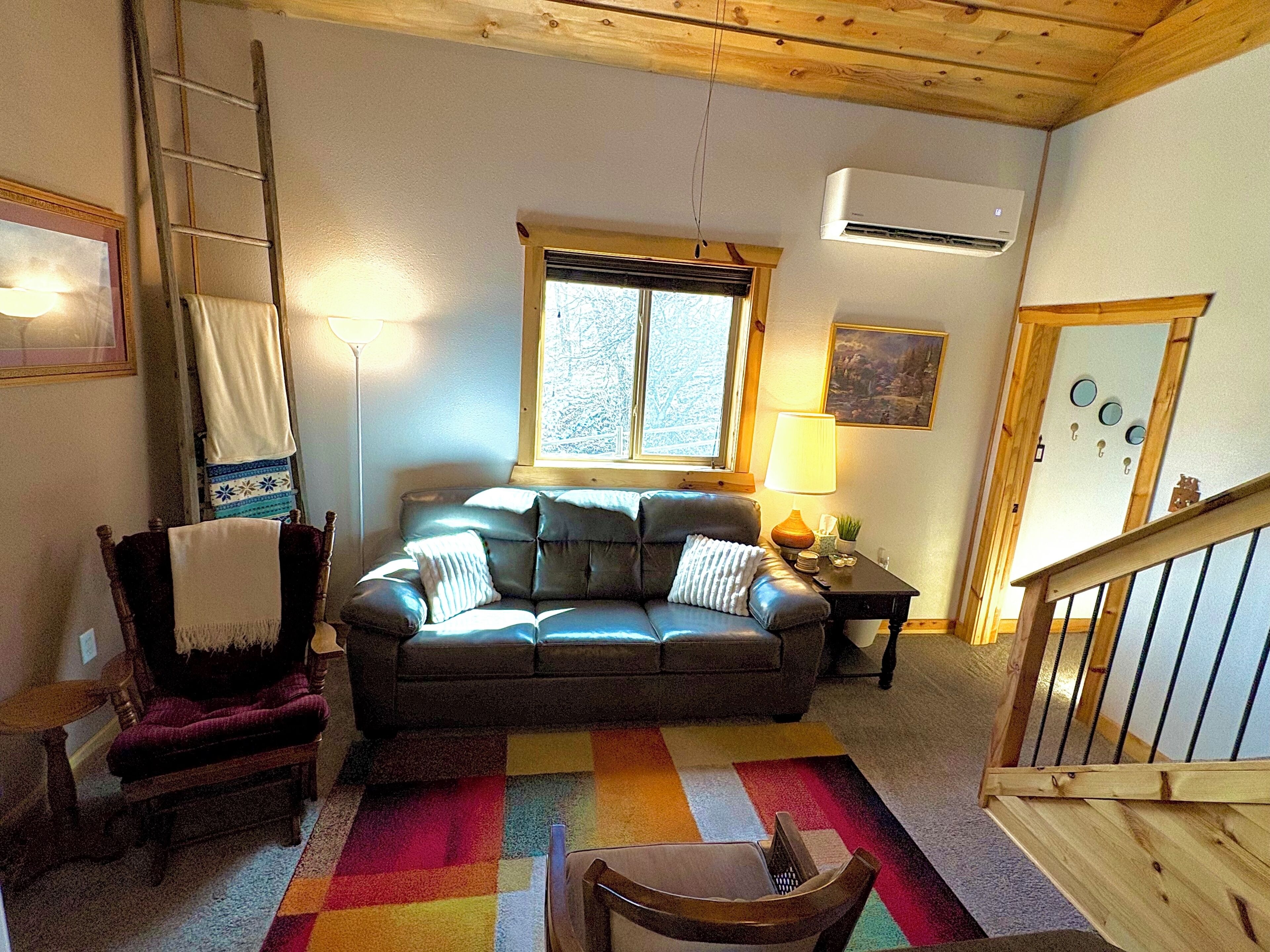 Un salon chaleureux avec un canapé en cuir, un fauteuil en bois, un tapis coloré, une fenêtre donnant sur un beau paysage et un escalier.