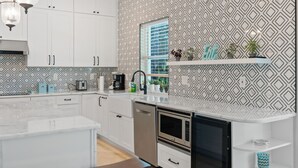 Cozinha moderna com armários brancos, lava-louças, micro-ondas e pia. Possui backsplash estampado e janela com persianas.