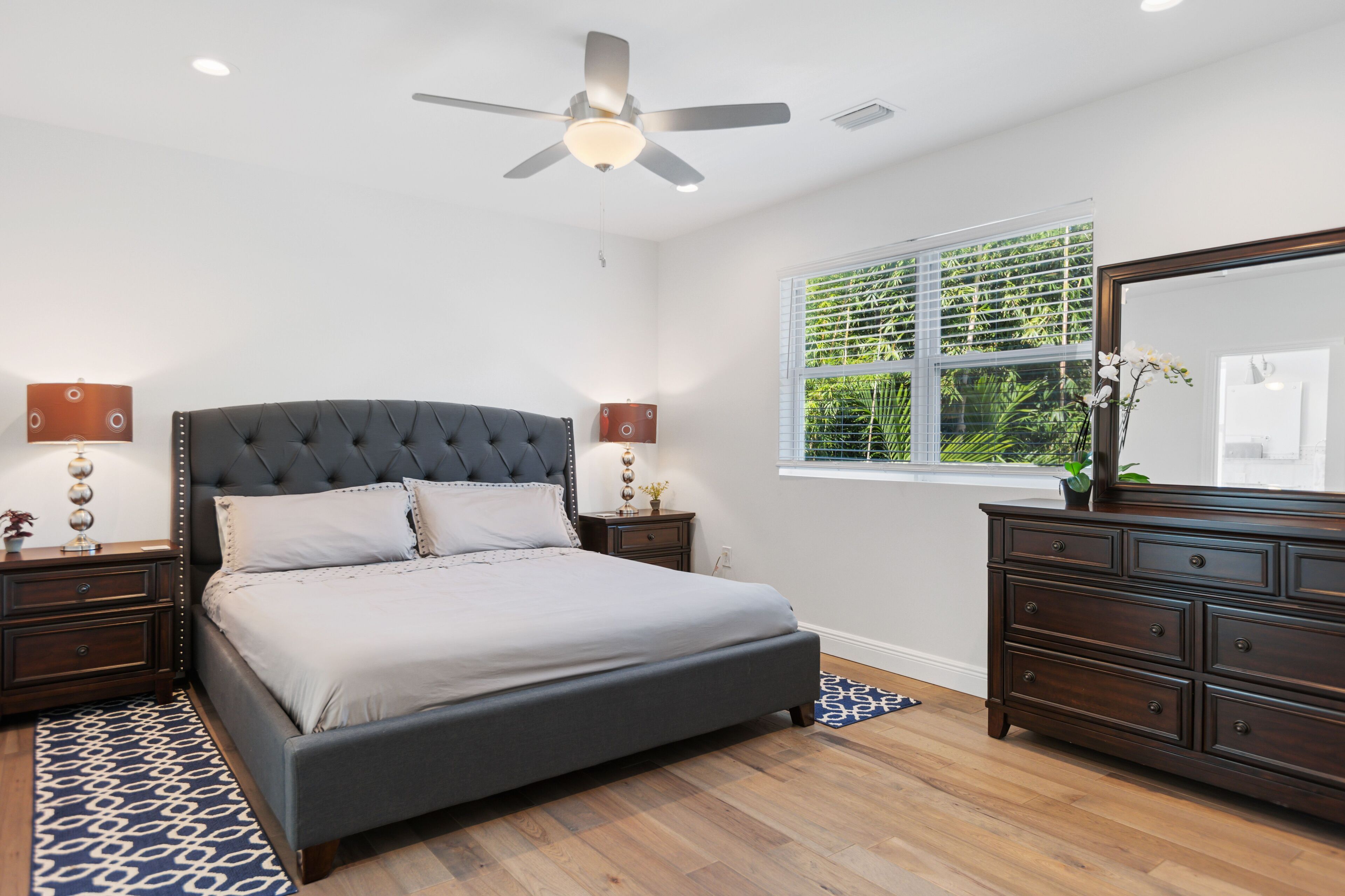 A bedroom with a large bed, a ceiling fan, a dresser, and a window with blinds.
