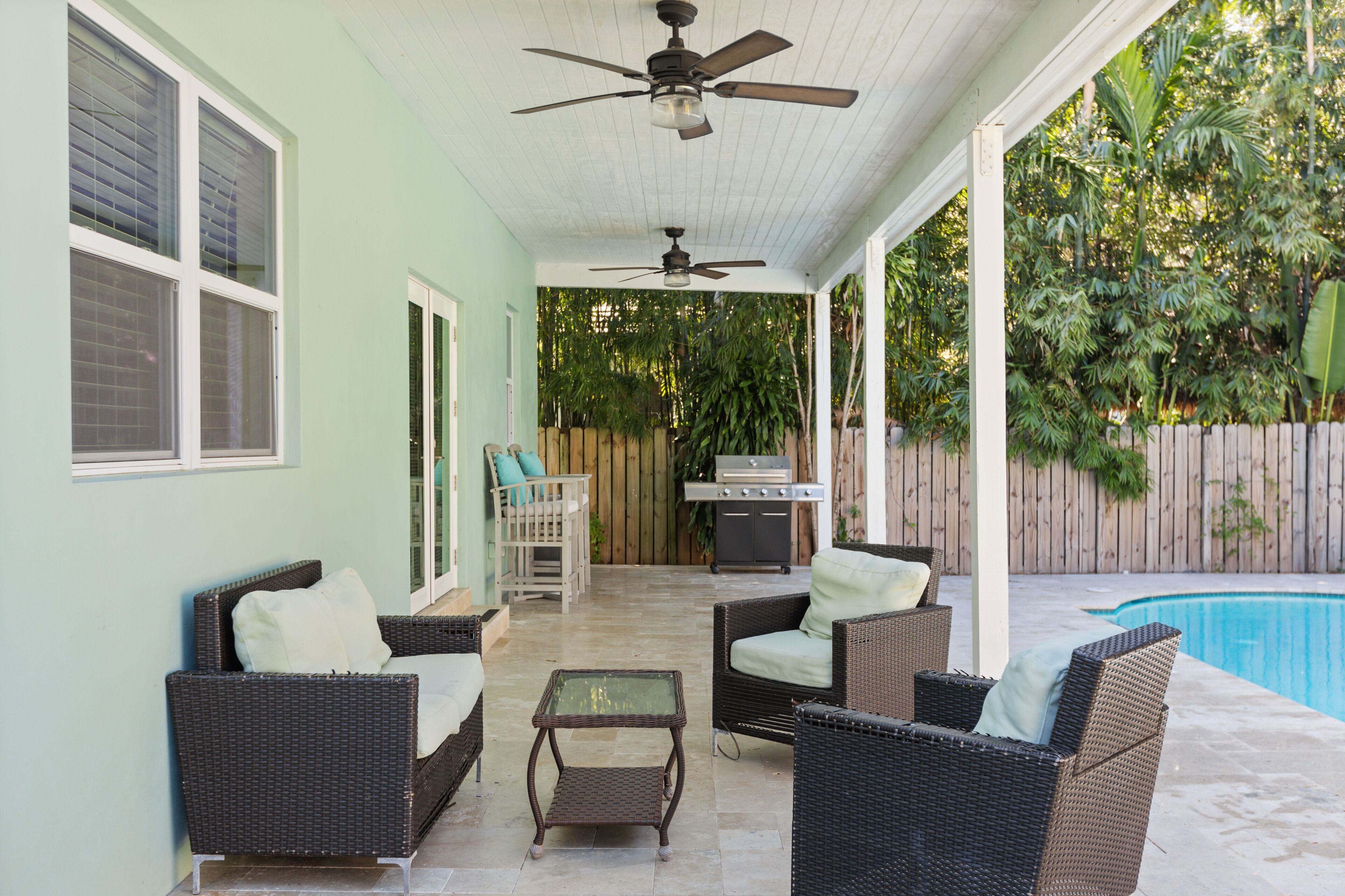 A covered patio with wicker furniture, a ceiling fan, and a pool in the background.