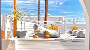 Un espacio al aire libre iluminado por el sol, con mesa y sillas de madera, plantas en macetas y vista al mar a través de un ventanal arqueado.