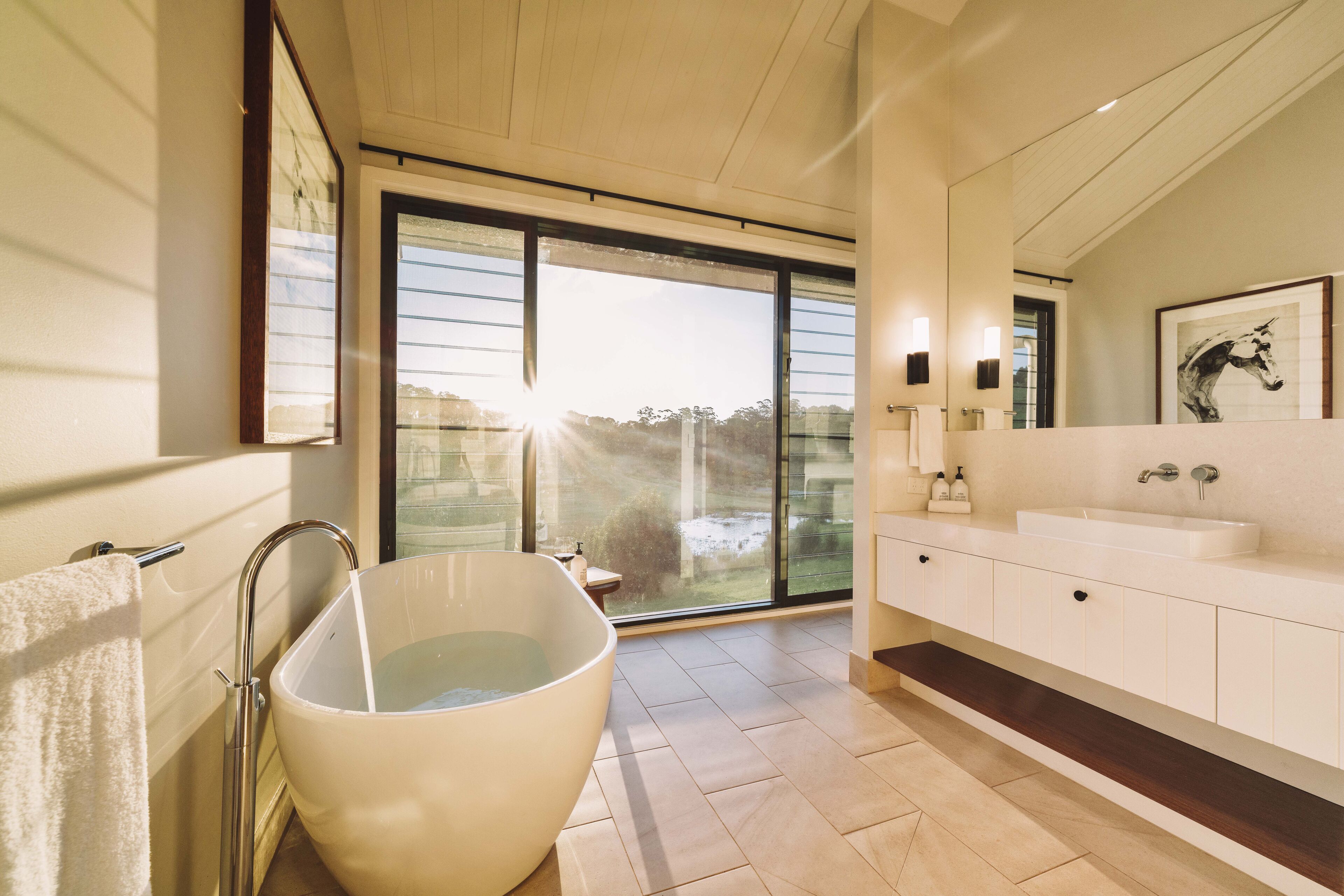 A modern bathroom with a large bathtub, a double sink vanity, and a large window providing a view of the outdoors.