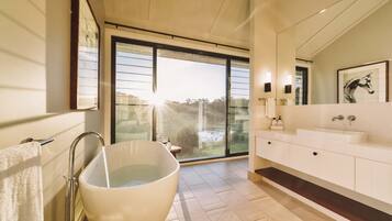 A modern bathroom with a large bathtub, a double sink vanity, and a large window providing a view of the outdoors.
