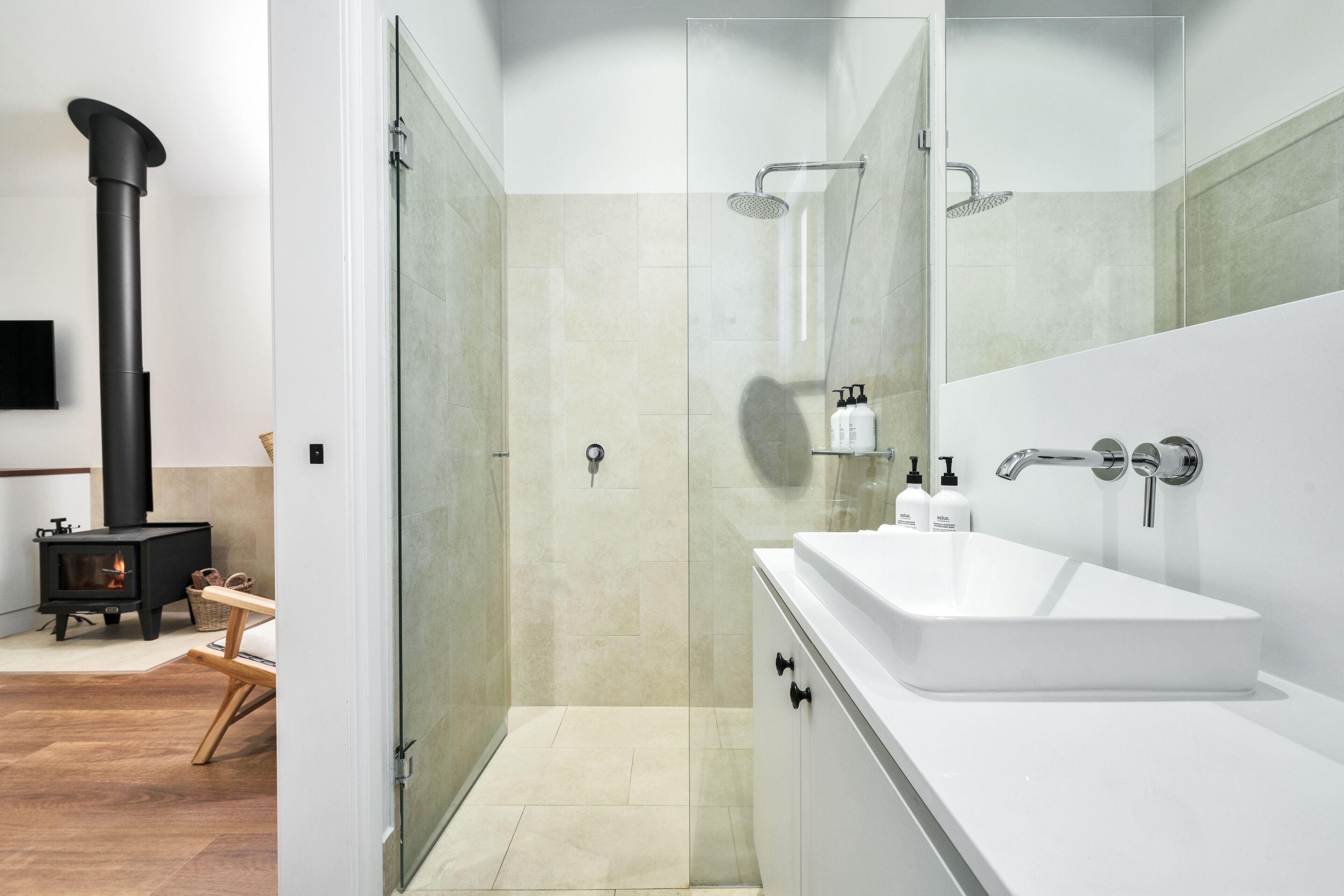 A modern bathroom with a white sink, glass shower enclosure, and a black wood-burning stove.