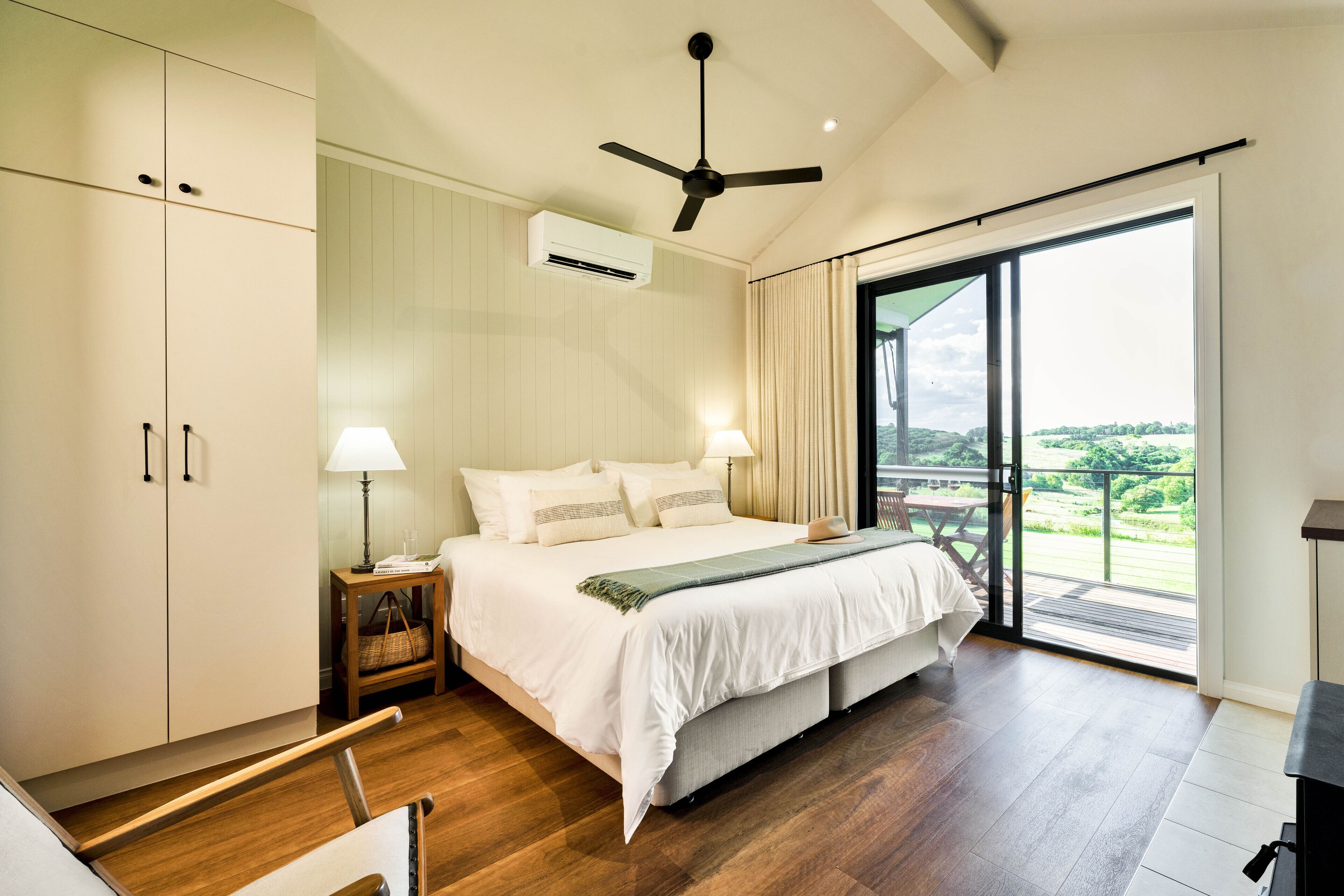 A bedroom with a large bed, a ceiling fan, an air conditioning unit, a nightstand with a lamp, and a view of a balcony.