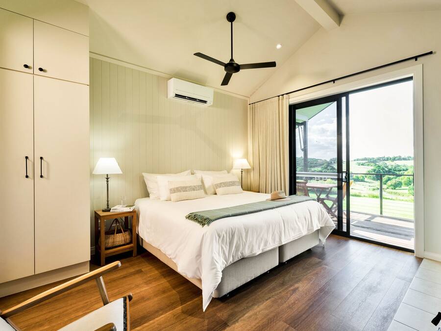 Un dormitorio con una cama grande, ventilador de techo, equipo de aire acondicionado, mesita de noche con lámpara y vistas a un balcón.