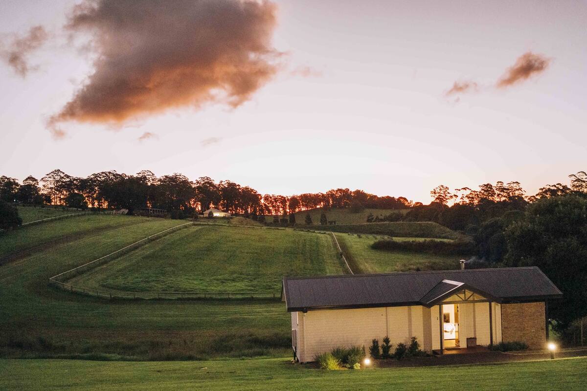 A small, single-story building with a sloped roof, situated in a grassy field with a backdrop of trees and hills.