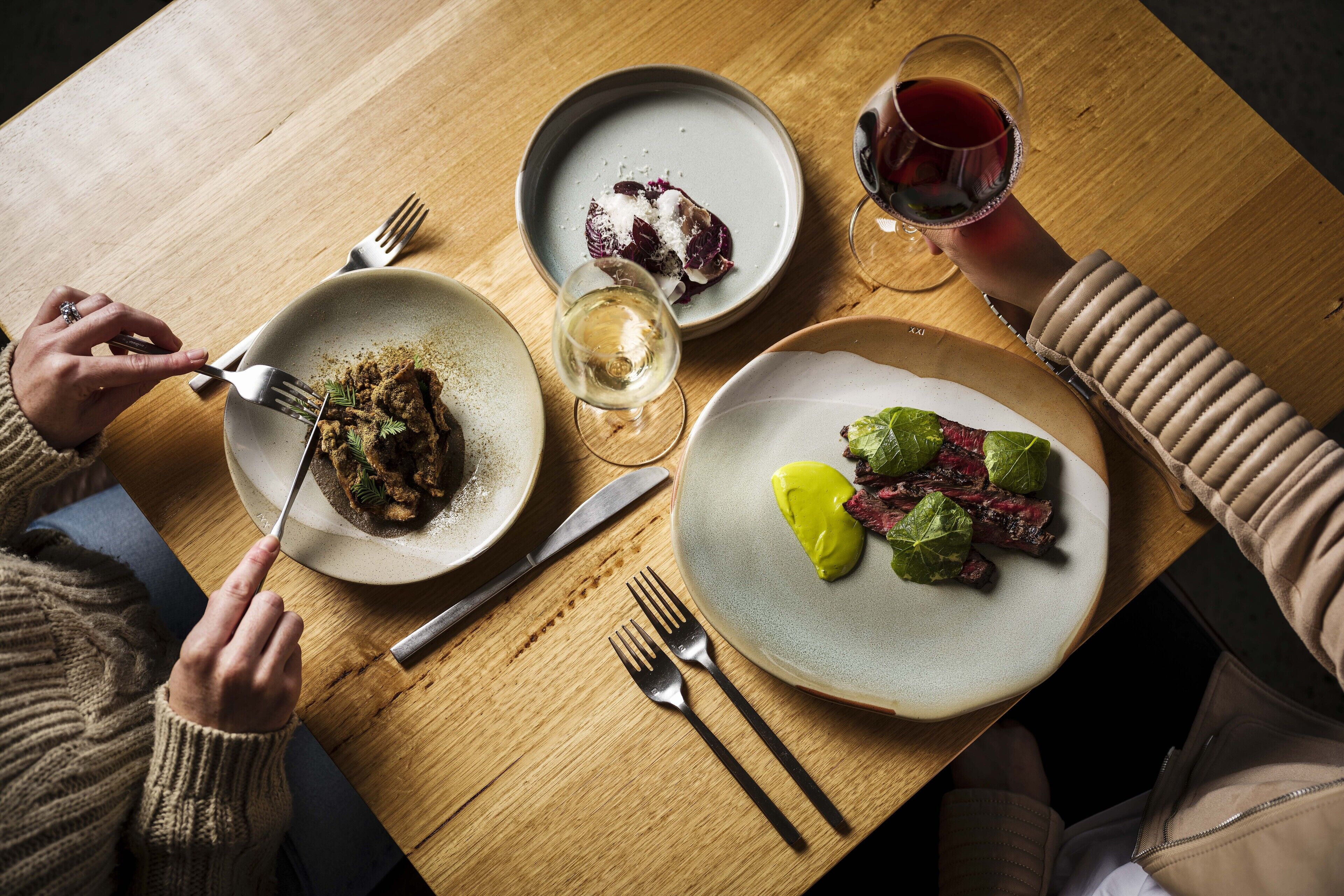 A wooden table with plates of food, a wine glass, and cutlery.