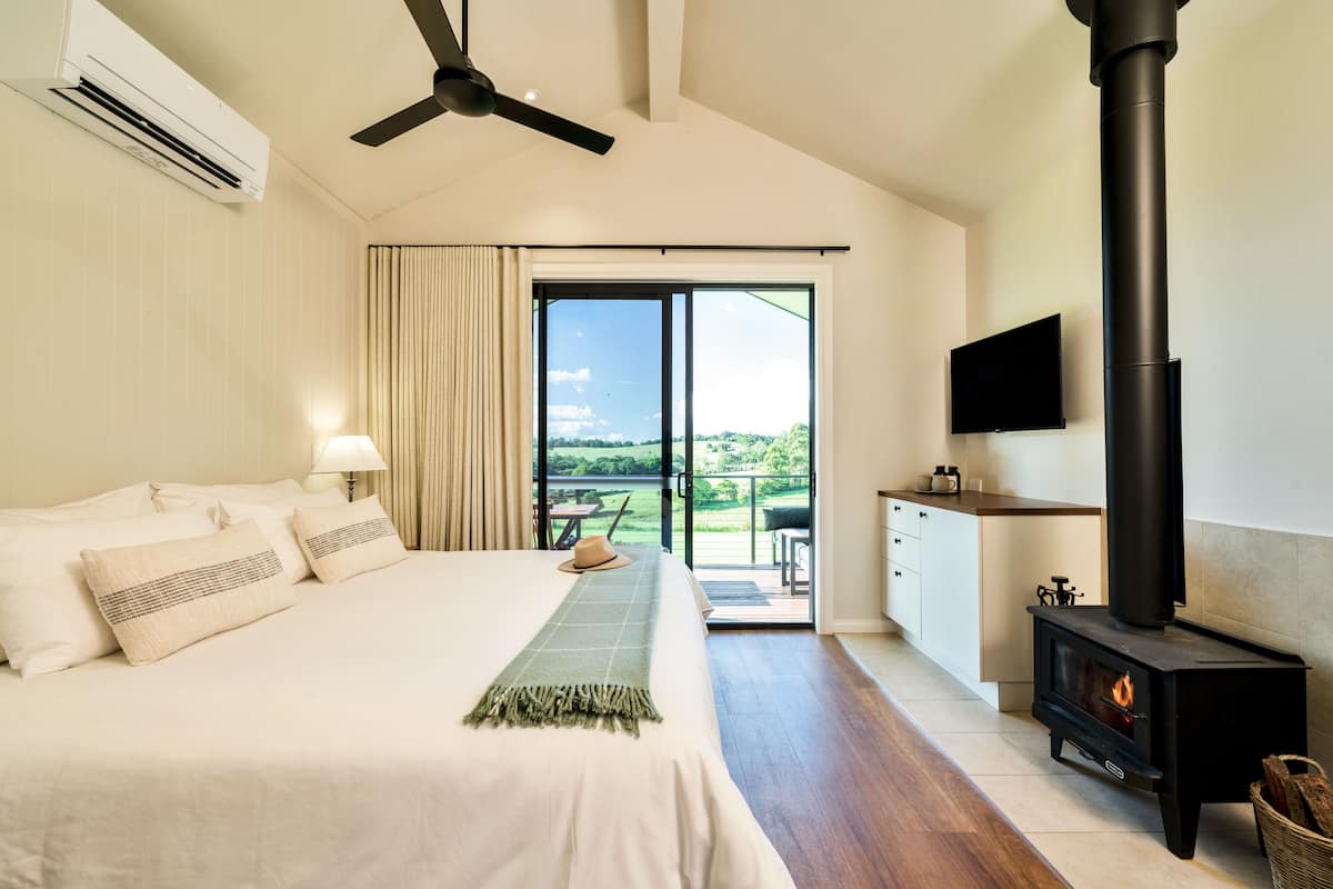 A bedroom with a bed, a ceiling fan, a fireplace, a TV, and a balcony with a view.