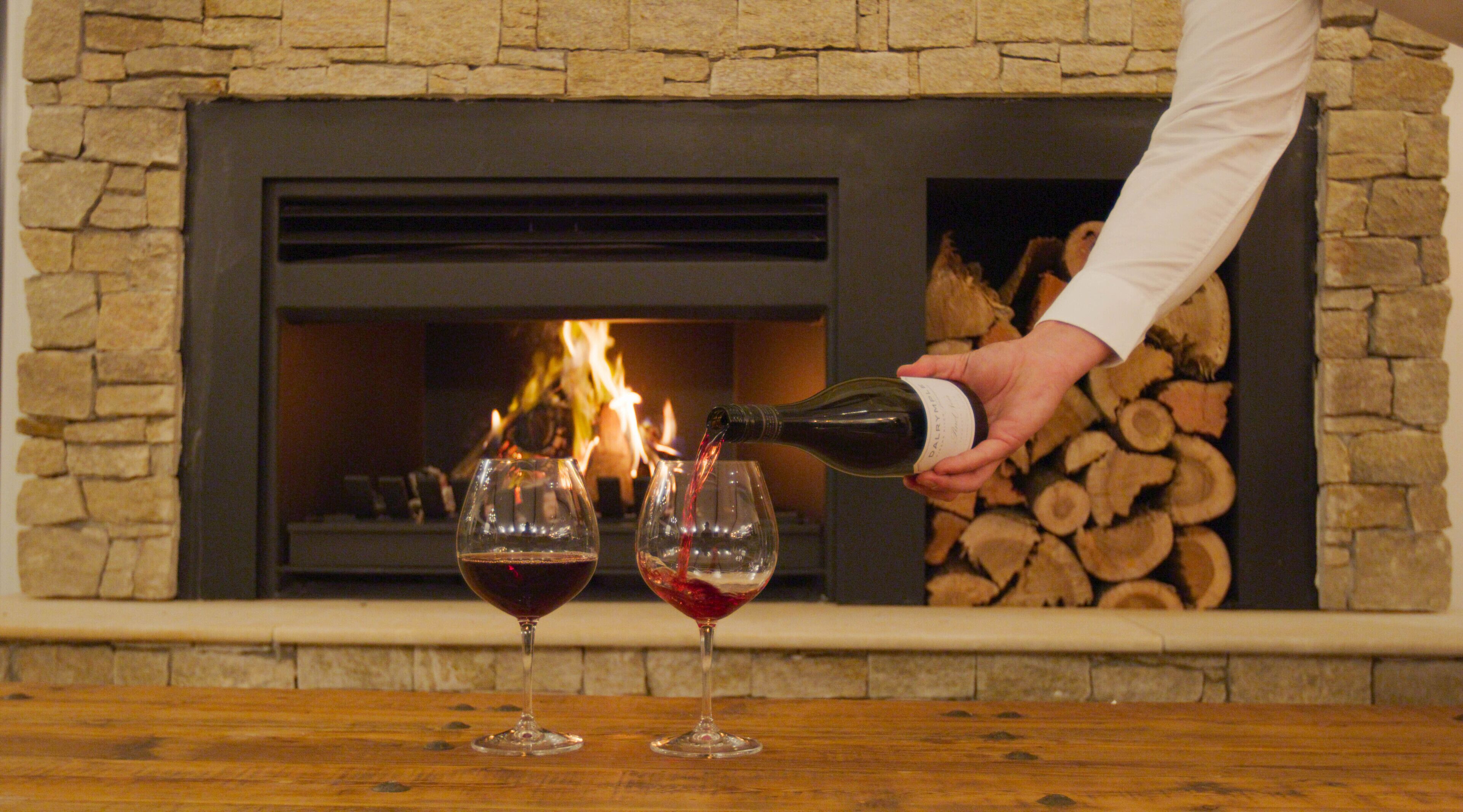 A person pouring wine into two glasses in front of a fireplace with logs.