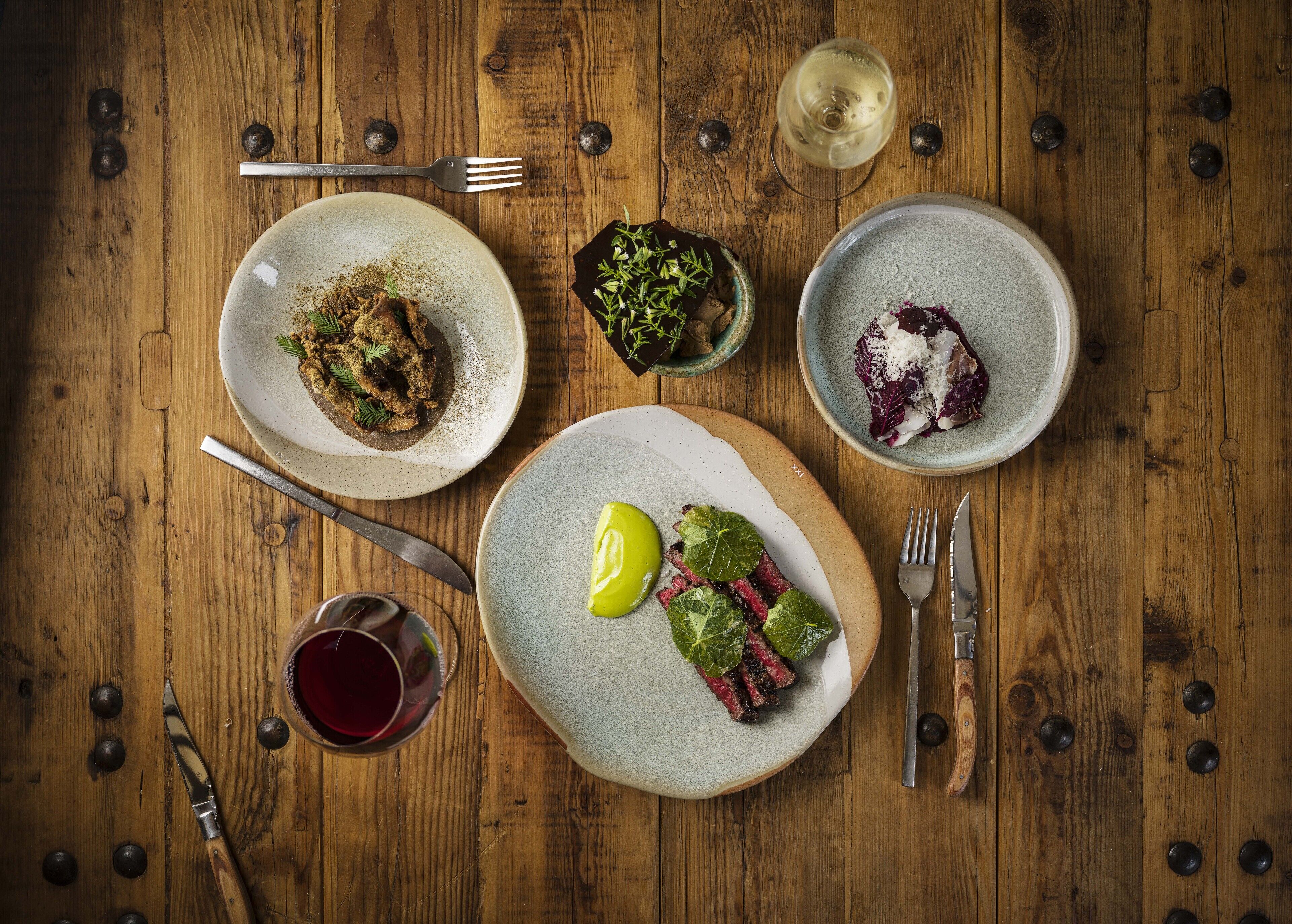 A wooden table with four plates of food, a glass of red wine, and cutlery.