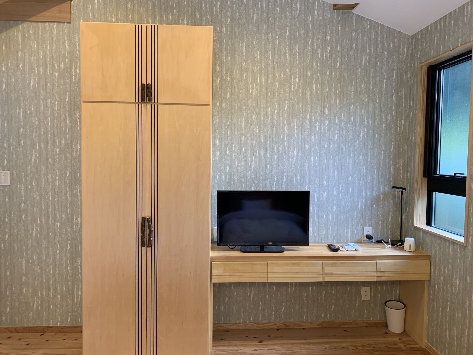 Une chambre avec une armoire en bois, un téléviseur posé sur un bureau en bois, une fenêtre et une poubelle.