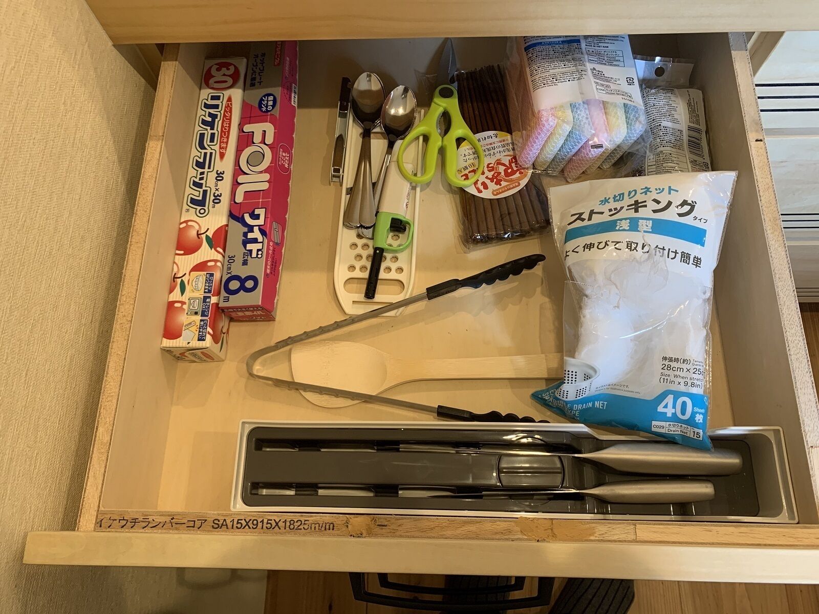 A drawer containing various kitchen utensils, including a pair of scissors, a spoon, a spatula, and a package of drain cleaner.