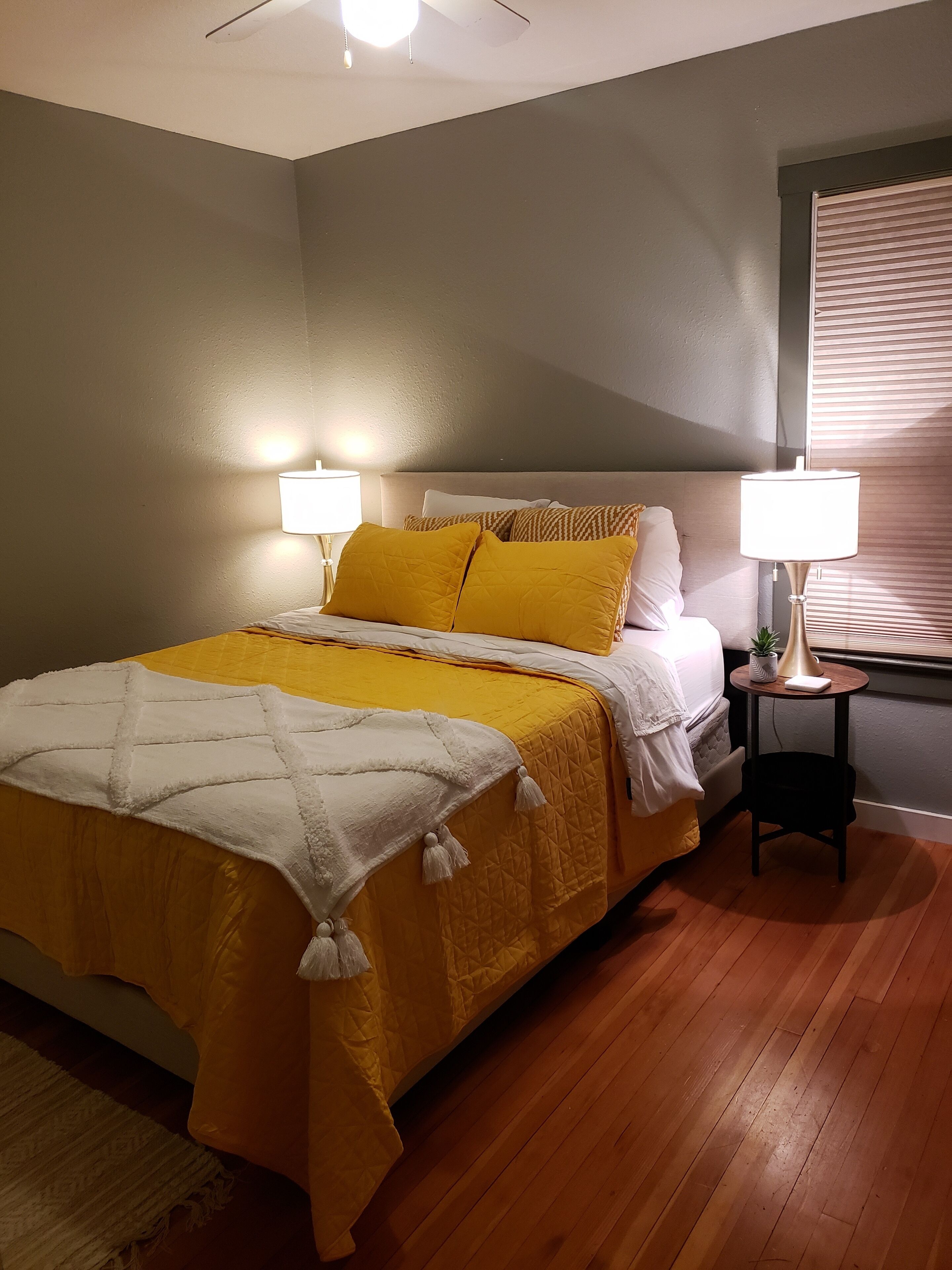 Ein Schlafzimmer mit einem Bett, zwei Nachttischlampen, einem kleinen Tisch mit Lampe, einer Zimmerpflanze und einem Fenster mit Jalousien.