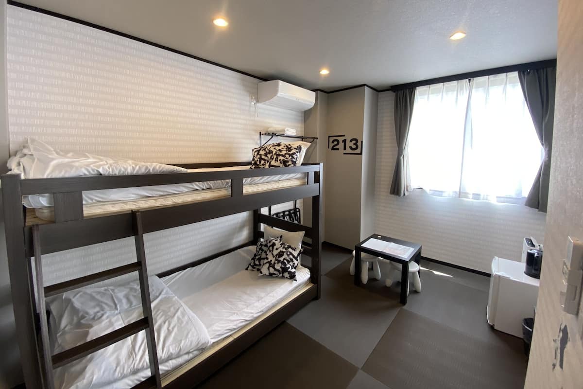 A bunk bed room with a small table and a desk, a window with curtains, and a wall number 213.