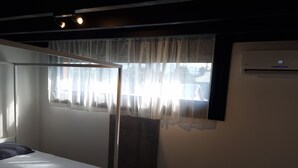A bedroom with a bed, a window with sheer curtains, and an air conditioning unit on the wall.