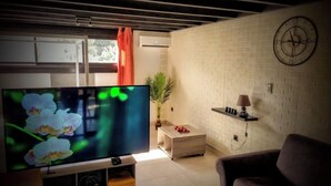 A living room with a flat-screen TV, a clock on the wall, a plant, and a small table with a lamp.