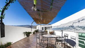 Un patio al aire libre con una mesa redonda y sillas, plantas en macetas y una pérgola con luces colgantes.