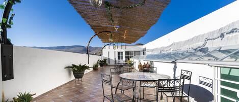 Un patio al aire libre con una mesa redonda y sillas, plantas en macetas y una pérgola con luces colgantes.