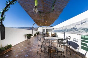 Un patio al aire libre con una mesa redonda y sillas, plantas en macetas y una pérgola con luces colgantes.