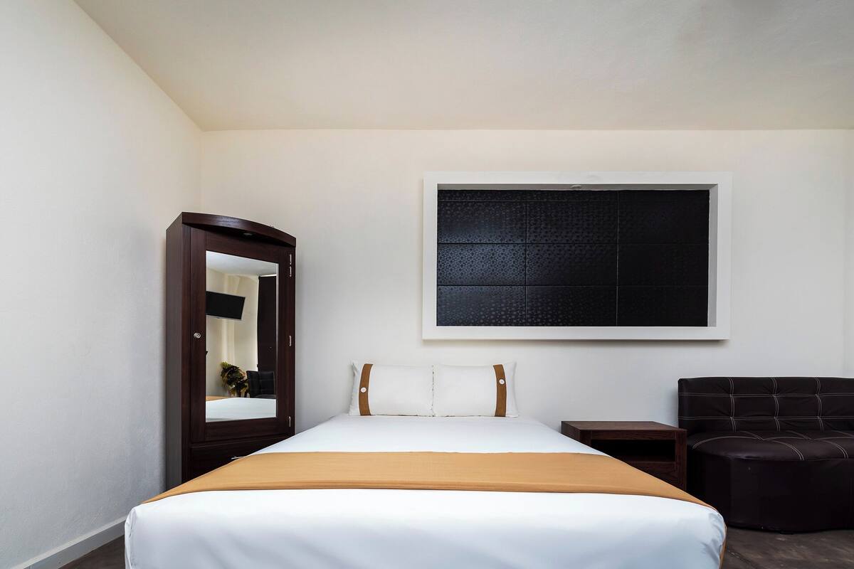 Une chambre d’hôtel avec un lit, un miroir, une petite table, une chaise et une grande fenêtre.
