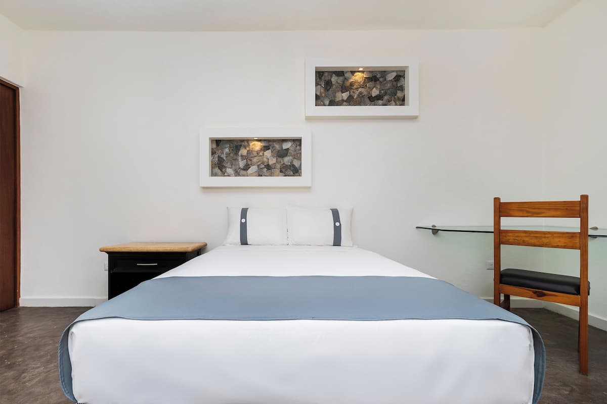 Une chambre à coucher comprenant un lit, une chaise en bois, une table de chevet et deux œuvres d’art encadrées accrochées au mur.