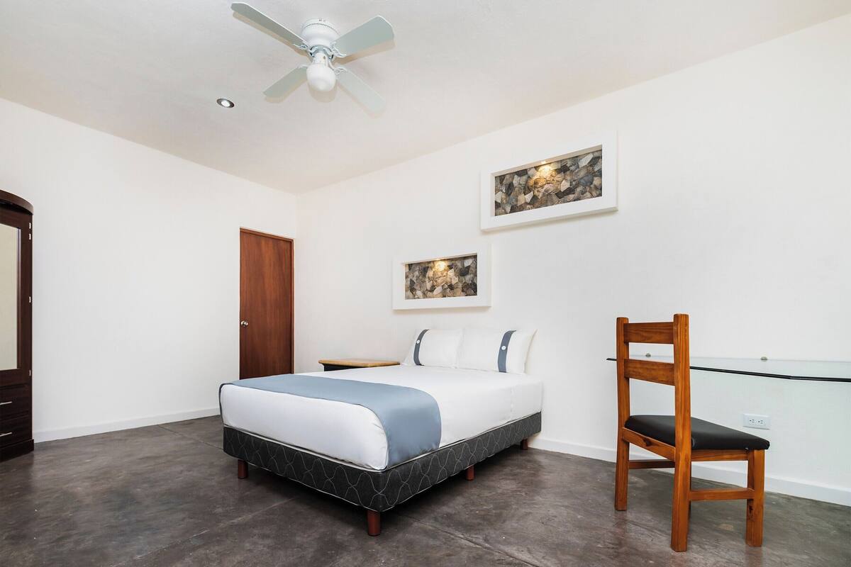 Une chambre à coucher avec un lit, une chaise en bois, un ventilateur de plafond et deux tableaux encadrés accrochés au mur.