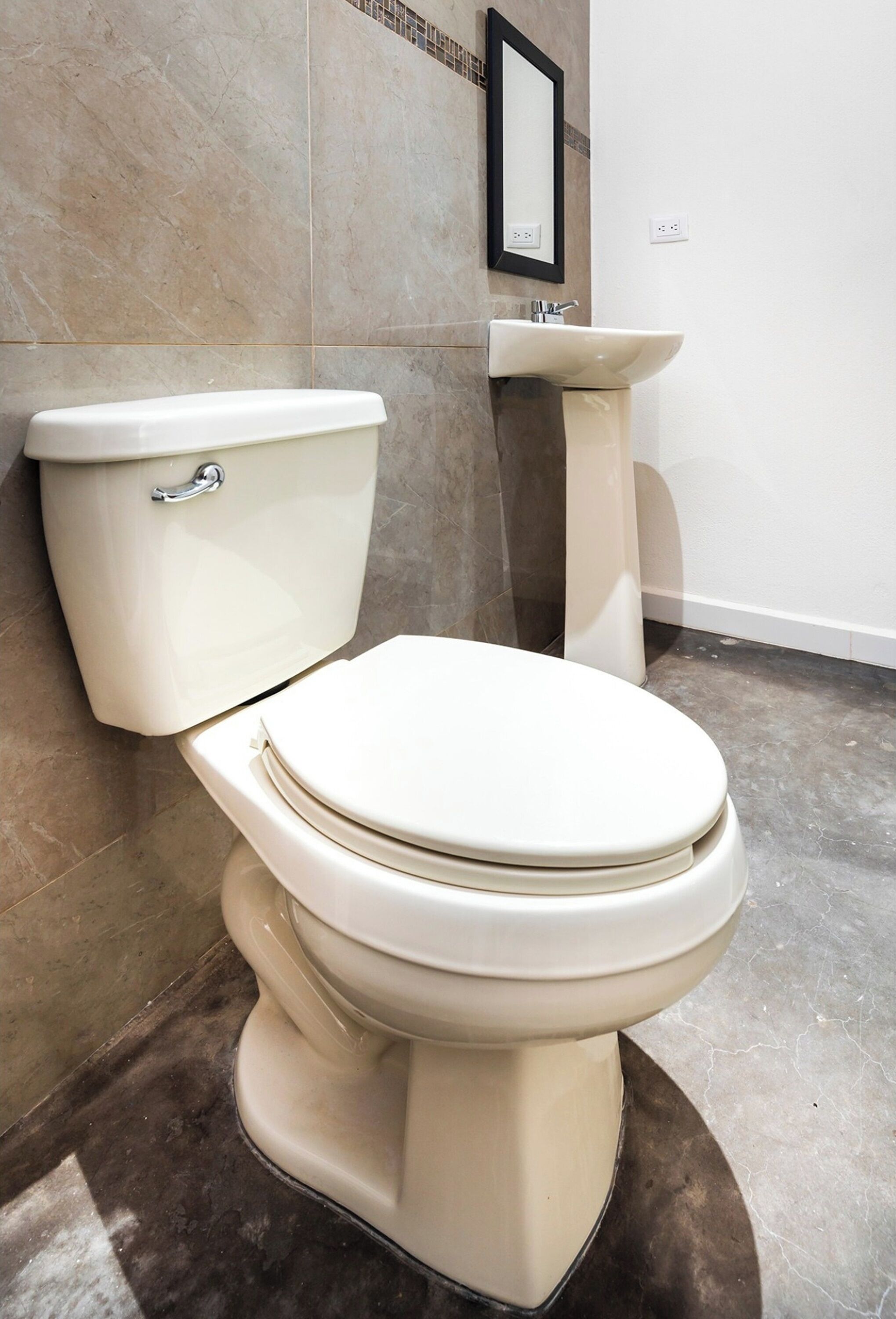 Een toilet met reservoir en zitje, een zuilwastafel en een betegelde muur met een inlijsting geplaatste spiegel.