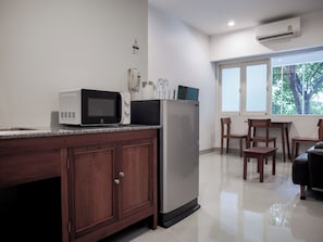 A kitchen with a microwave, refrigerator, and a coffee maker on the counter. There are wooden cabinets and a window with a view of trees. - Niagara Hotel (Bangkok)
