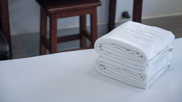 A stack of white towels on a white surface, a wooden chair, and a wooden stool in a room. - Niagara Hotel (Bangkok)