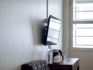 A wall-mounted television, a water bottle, a coffee maker, and a small table with a cushion in a room with a window. - Niagara Hotel (Bang Rak)