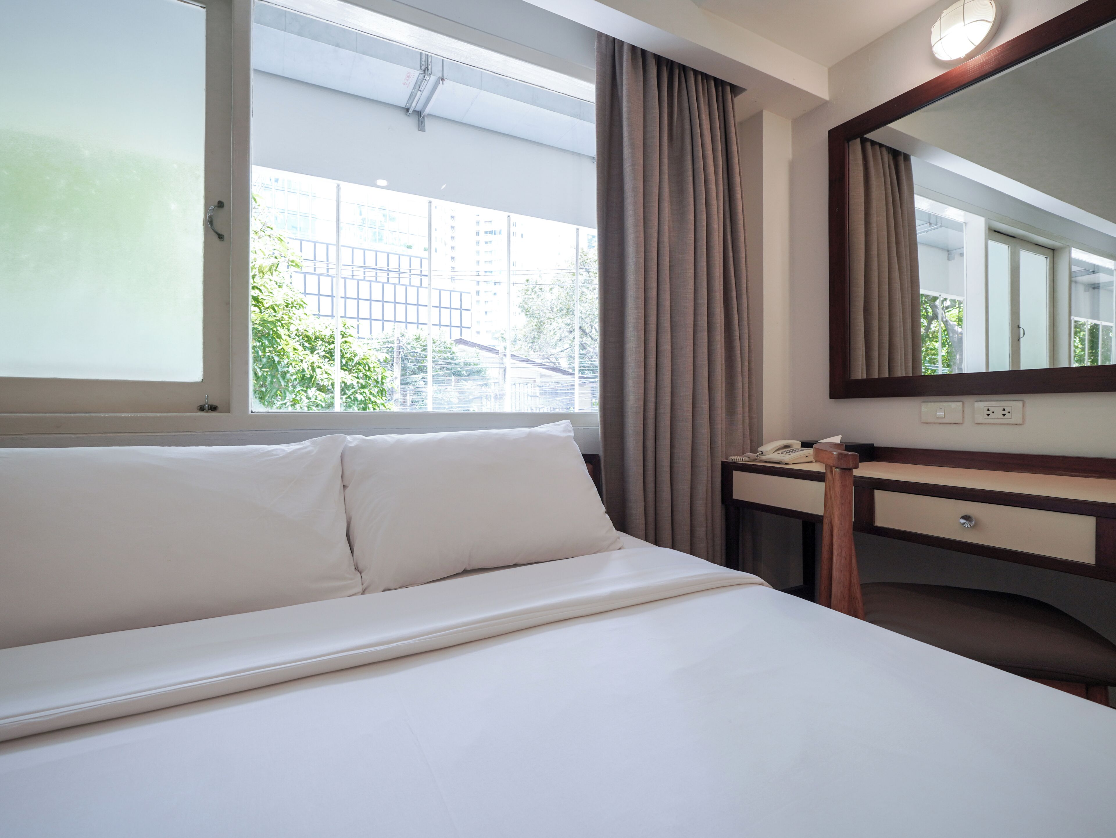 A hotel room with a bed, a bedside table with a telephone, a mirror, and a window with curtains.