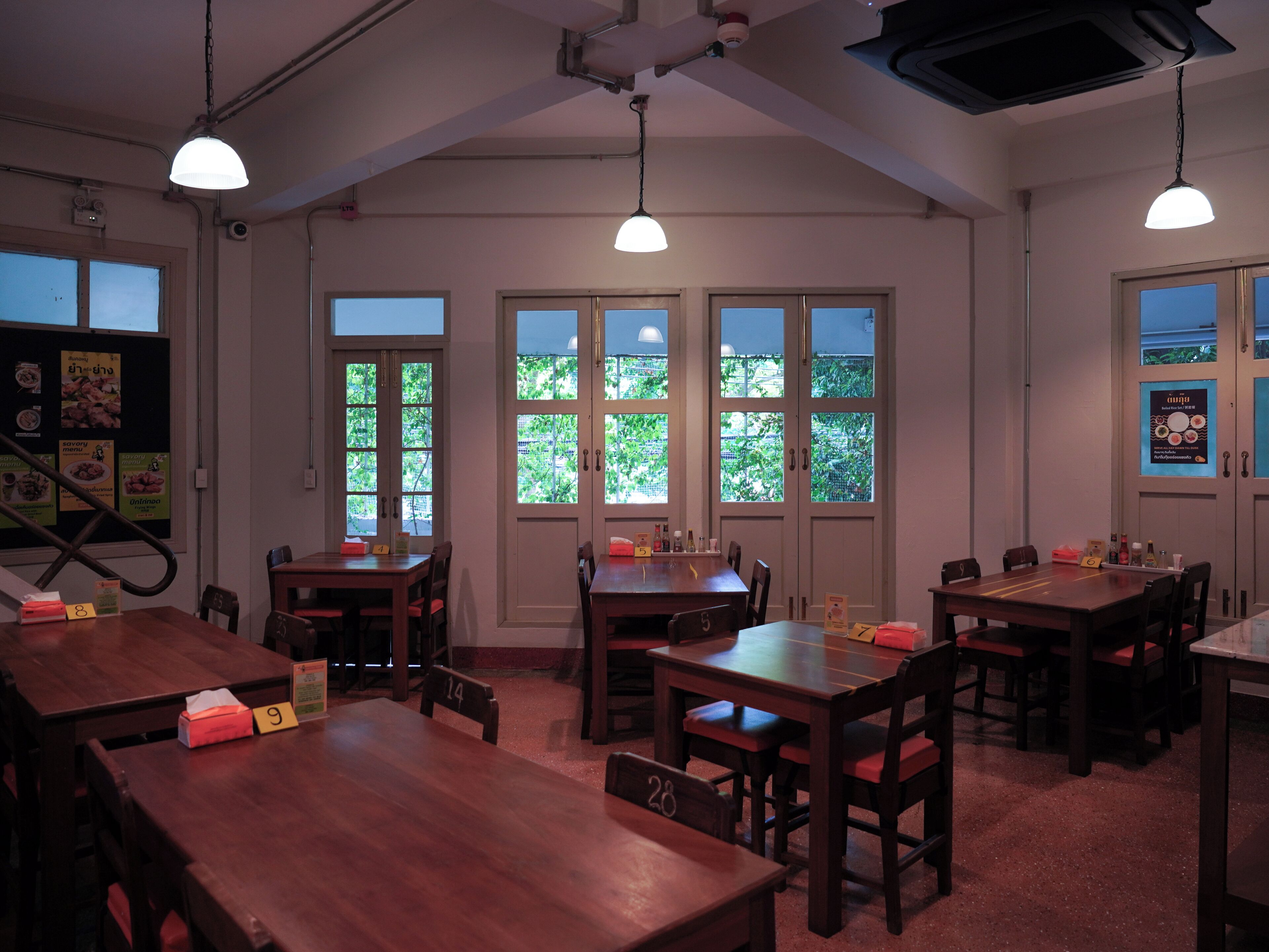 Interior de restaurante con varias mesas y sillas de madera, ventanas amplias con follaje exterior e iluminación colgante.