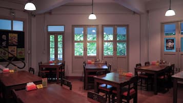 Interior de restaurante con varias mesas y sillas de madera, ventanas amplias con follaje exterior e iluminación colgante.