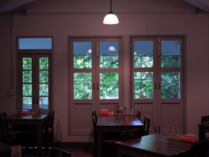 A dining area with a table and chairs, a hanging light fixture, and windows with a view of greenery. - Niagara Hotel (Bangkok)